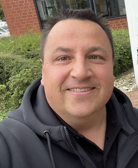 Mann in schwarzem Hoodie lächelt in die Kamera, im Hintergrund sind Büsche und ein Gebäude mit roten Klinkern zu sehen.